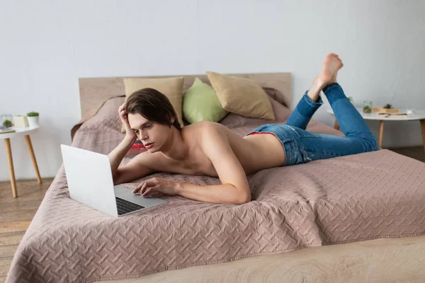 Hemdloser transsexueller junger Mann in Jeans mit Laptop auf dem Bett — Stockfoto