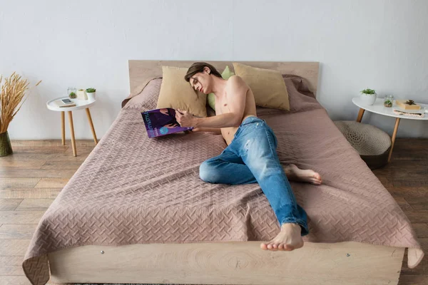 Shirtless transgender young man in jeans reading magazine on bed — Stock Photo