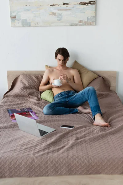 Torse nu transgenre jeune homme en jeans tenant tasse de café tout en regardant un film sur ordinateur portable dans la chambre — Photo de stock