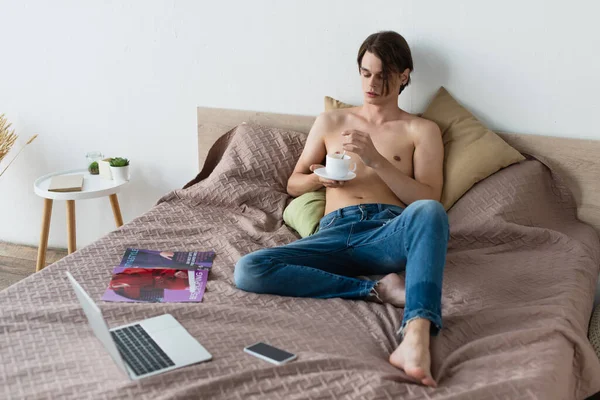 Barefoot transgender young man in jeans holding cup of coffee near gadgets on bed — Stock Photo