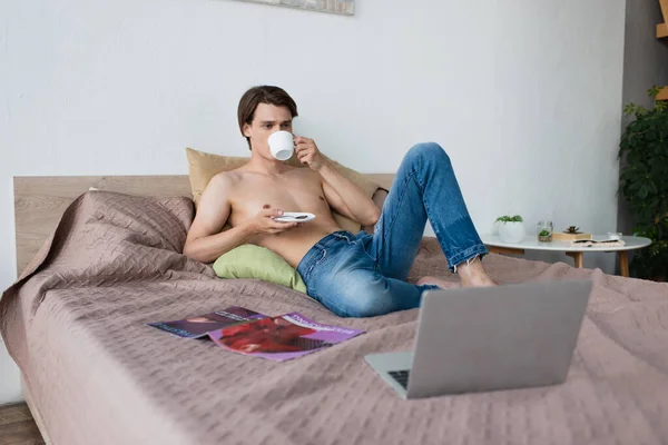 Безрубашечный трансгендерный молодой человек в джинсах держит чашку и пьет кофе возле ноутбука на кровати — стоковое фото