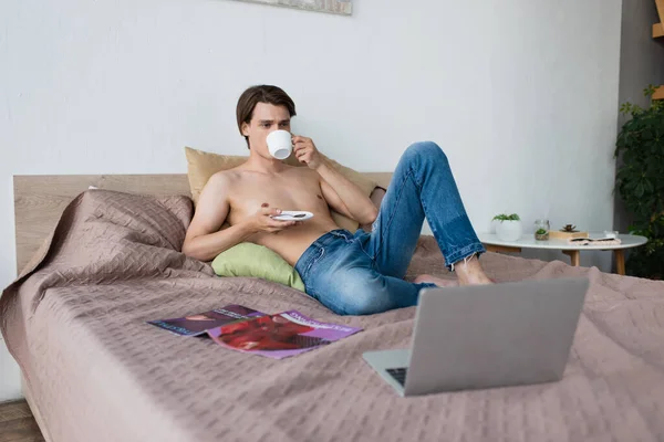 Senza maglietta transgender giovane in jeans in possesso di tazza e bere caffè vicino computer portatile sul letto — Foto stock