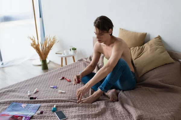 Joven transgénero en jeans sosteniendo esmalte de uñas en el dormitorio - foto de stock