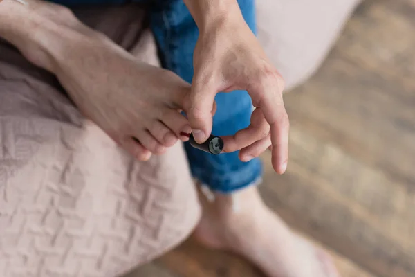 Vue recadrée du jeune homme transgenre en jeans tenant du vernis à ongles et faisant pédicure dans la chambre — Photo de stock