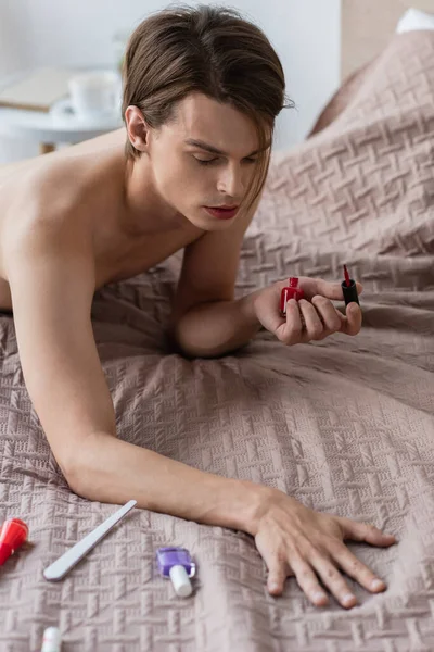 Hemdloser Transgender-junger Mann in Jeans hält Nagellack in der Hand und blickt im Schlafzimmer auf die Hand — Stockfoto