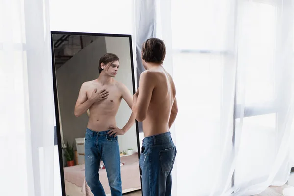 Hemdloser Transgender-junger Mann in Jeans steht mit der Hand auf der Hüfte vor dem Spiegel — Stockfoto