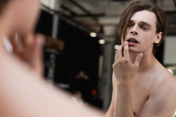 Sin camisa transgénero joven tocando el labio y mirando el espejo - foto de stock