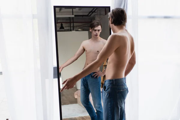 Без сорочки транссексуал молодий чоловік в джинсах стоїть з рукою на стегні і дивиться на дзеркало — стокове фото