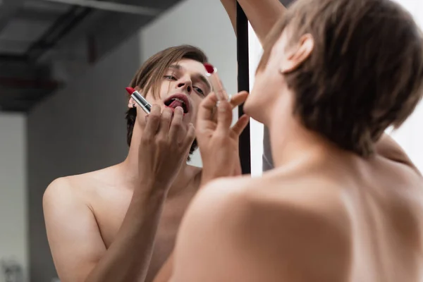 Hemdloser Transgender-junger Mann trägt Lippenstift auf, während er in den Spiegel schaut — Stockfoto