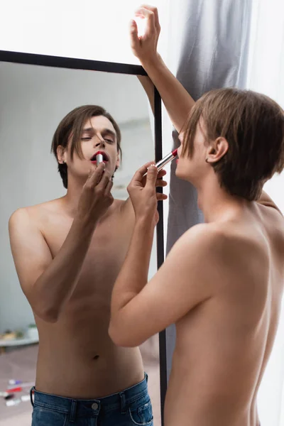 Torse nu transgenre jeune homme appliquant rouge à lèvres et regarder miroir à la maison — Photo de stock