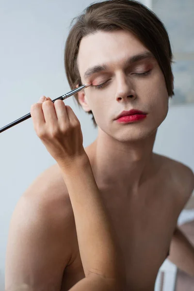 Maquillador aplicando sombra de ojos con pincel cosmético sobre hombre transgénero - foto de stock