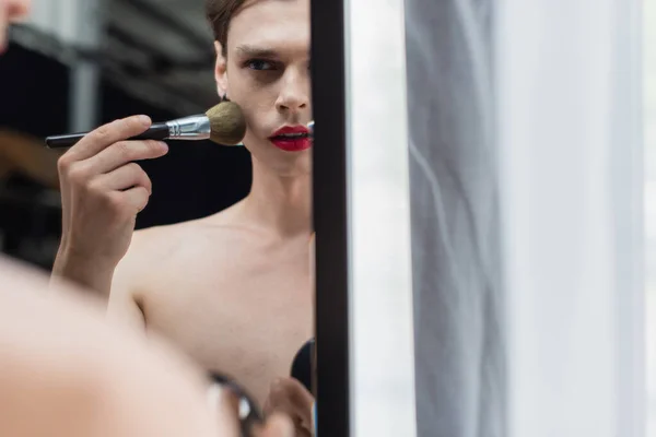 Jeune homme transgenre appliquant de la poudre visage avec pinceau cosmétique près du miroir — Photo de stock
