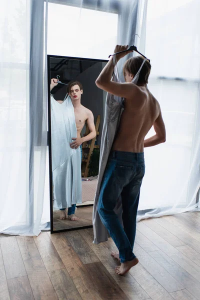 Pleine longueur de jeune homme transgenre avec maquillage tenant cintre avec robe glissante près du miroir — Photo de stock
