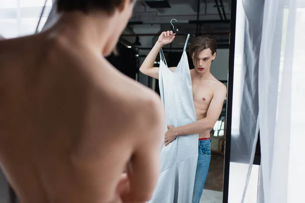Young transgender man with makeup holding hanger with slip dress and looking at mirror — Stock Photo