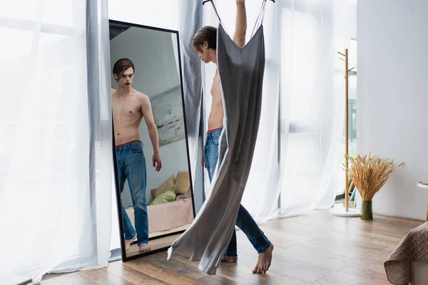 Comprimento total do homem transgênero sem camisa com maquiagem segurando cabide com vestido deslizante perto do espelho em casa — Fotografia de Stock