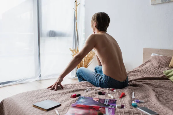 Vue arrière de l'homme transgenre torse nu assis sur le lit près des produits de maquillage — Photo de stock