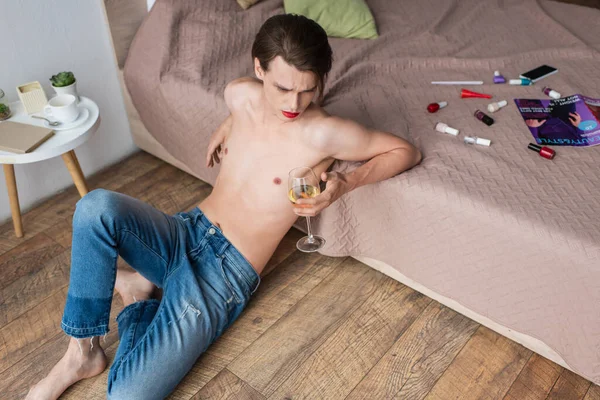 Vue grand angle de jeune homme transgenre tenant un verre de vin tout en étant assis près du lit avec des produits de maquillage — Photo de stock