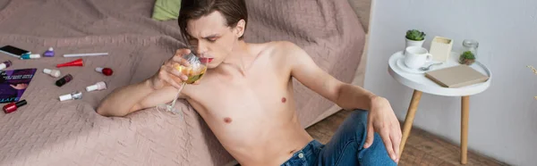 Jeune homme transgenre boire du vin tout en étant assis près du lit avec des produits de maquillage, bannière — Photo de stock