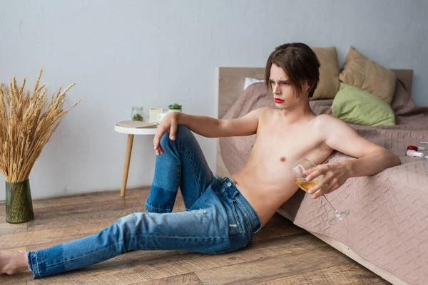 Frustré jeune homme transgenre tenant un verre de vin tout en étant assis près du lit — Photo de stock