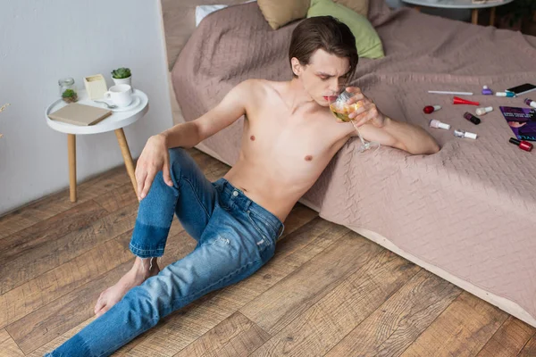 Junger Transgender-Mann trinkt Wein, während er neben dem Bett mit Schminkprodukten sitzt — Stockfoto