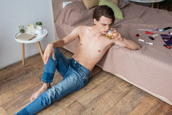Young transgender man drinking wine while sitting near bed with makeup products — Stock Photo