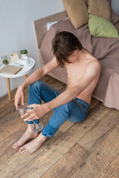 Vista de ángulo alto de triste joven transexual sosteniendo un vaso de vino mientras está sentado cerca de la cama - foto de stock