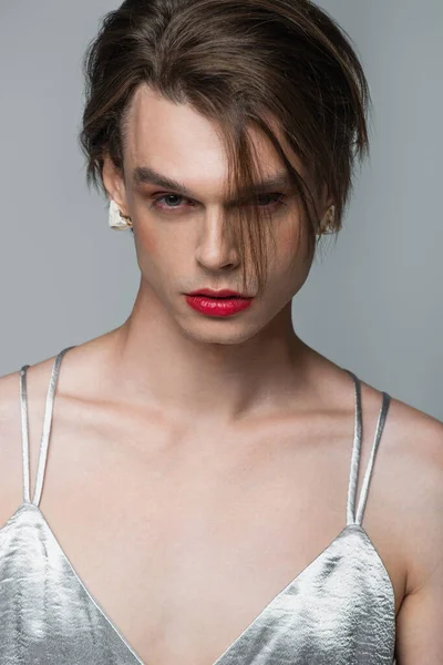 Junger Transgender-Mann mit Make-up und Ohrring blickt isoliert in die Kamera auf grau — Stockfoto