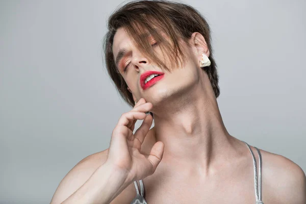 Joven hombre transgénero con maquillaje y pendiente posando con los ojos cerrados aislados en gris - foto de stock