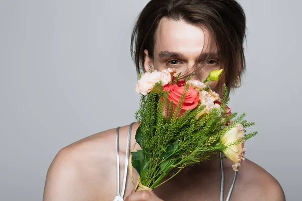 Joven transgénero hombre sosteniendo flores mientras cubre la cara aislado en gris - foto de stock