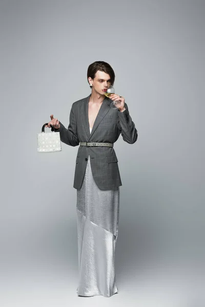 Full length of transgender man in slip dress and blazer drinking wine while holding purse on gray — Stock Photo