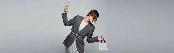 Homme transgenre en slip robe avec blazer tenant sac à main et verre de vin isolé sur gris, bannière — Photo de stock