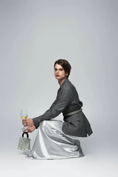 Transgender-Mann in voller Länge im Slip-Kleid mit Blazer, der Handtasche und Glas Wein hält, während er auf grau sitzt — Stockfoto