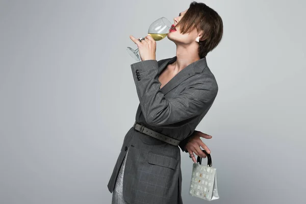 Hombre transgénero en chaqueta bebiendo vino mientras sostiene bolso aislado en gris - foto de stock