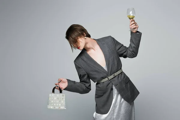 Young transgender man in slip dress with blazer holding purse and glass of wine isolated on grey — Stock Photo