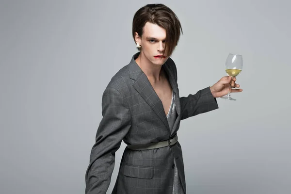 Young transgender man in blazer biting lip while holding glass of wine isolated on grey — Stock Photo