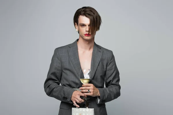 Jeune homme transgenre en blazer tenant sac à main et verre de vin isolé sur gris — Photo de stock