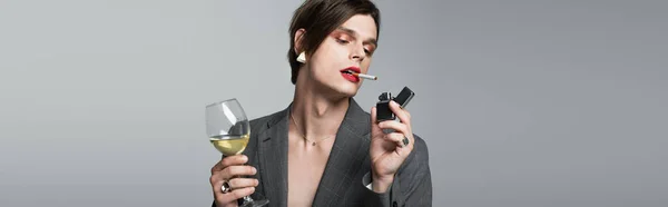 Молодой трансгендер в пиджаке держа бокал вина и зажигалки во время курения изолированы на сером, баннер — стоковое фото