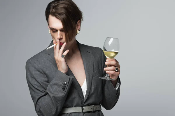 Junger Transgender-Mann im Blazer hält Glas Wein in der Hand und raucht isoliert auf Grau — Stockfoto