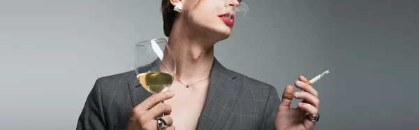Abgeschnittene Ansicht eines jungen Transgender-Mannes in Blazer und Ohrring, der ein Glas Wein hält, während er auf grau raucht, Banner — Stockfoto