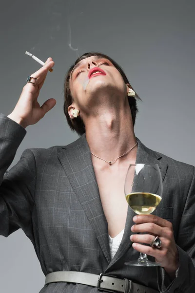 Jeune homme transsexuel en blazer et boucles d'oreilles tenant un verre de vin tout en fumant isolé sur gris — Photo de stock