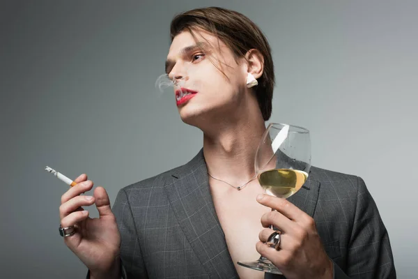 Молодой транссексуал в блейзере держит бокал вина и курение сигареты изолированы на серый — стоковое фото