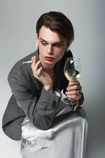 Junger transsexueller Mann im Blazer und Slip-Kleid hält ein Glas Wein in der Hand, während er Zigarette auf grau raucht — Stockfoto