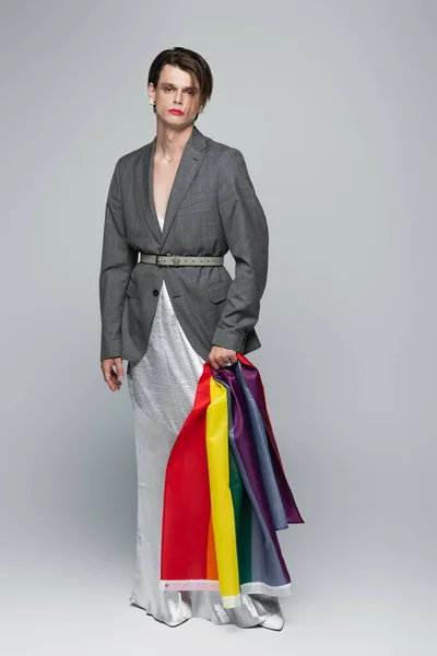 Full length of young transsexual man in blazer and slip dress holding lgbt flag on grey — Stock Photo