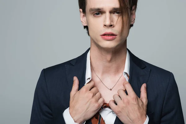 Joven transexual hombre de negocios en traje ajustando camisa y mostrando collar aislado en gris - foto de stock