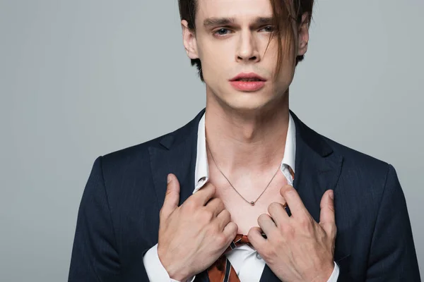 Young transsexual businessman in suit adjusting shirt and showing necklace isolated on grey — Stock Photo