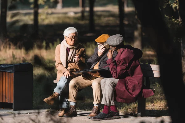 Hombres mayores multiétnicos con álbum de fotos sentados en el banco en el parque - foto de stock