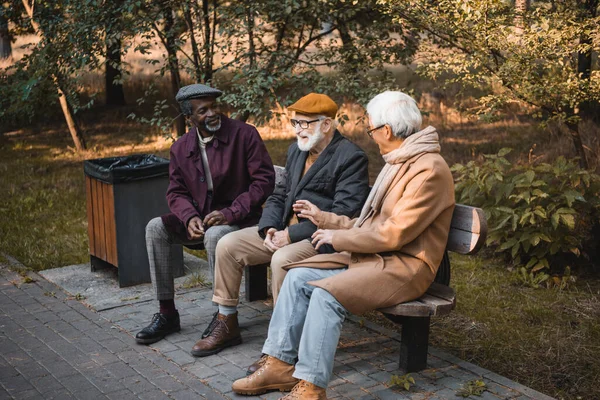 Sorrindo homens multiétnicos falando no banco no parque de outono — Fotografia de Stock