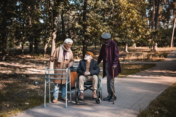 Amigos multiculturales con marco para caminar y silla de ruedas hablando en el parque de otoño - foto de stock