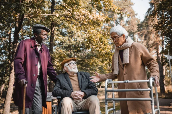 Ásia homem com andar quadro falando para multiétnico amigos com cadeira de rodas no parque — Fotografia de Stock