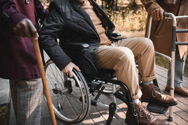 Обрезанный вид человека в инвалидной коляске рядом с межрасовыми друзьями с ходячей рамой и тростью в парке — стоковое фото